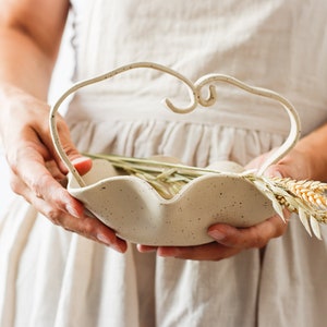 A woman in a beige apron holds a mottled beige ceramic basket in the shape of waves. It has a handle formed by two stoneware strips of the same color as the basket, which are joined in the center forming two small spirals. Contains spikes.