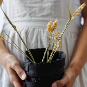 Two woman's hands hold a pot that looks out of focus. It is made from hand-cut strips of black clay with lots of texture. It contains some decorative dowels.