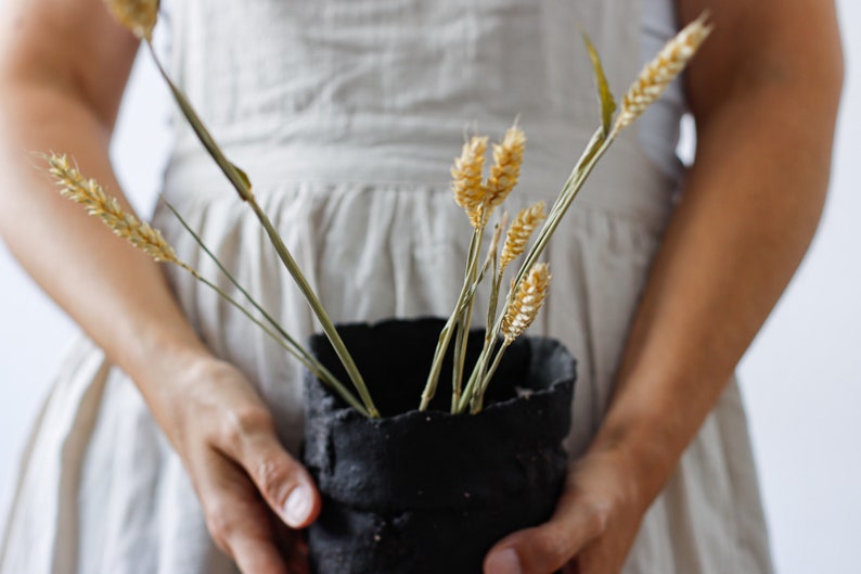 Two woman's hands hold a pot that looks out of focus. It is made from hand-cut strips of black clay with lots of texture. It contains some decorative dowels.