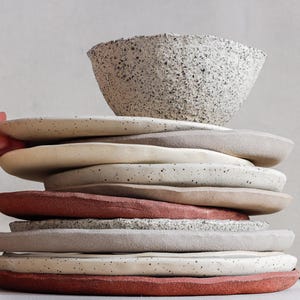 May include: A stack of handmade ceramic plates and a bowl in various neutral colors. The plates are stacked with a bowl on top, and the colors include speckled white, beige, and terracotta. The bowl and plates have a textured surface.