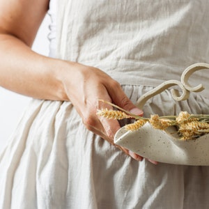 A woman in a beige apron holds a matte beige ceramic basket. Only a part of it is visible from the front. The basket is wavy and the handle can be seen, formed by two strips that join in the center forming two small spirals.