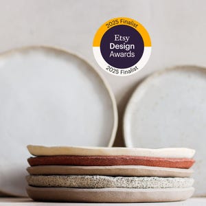 May include: A selection of handmade ceramic plates in cream, red, and speckled grey, with two larger white plates behind. The image features an Etsy Design Awards 2025 Finalist badge.