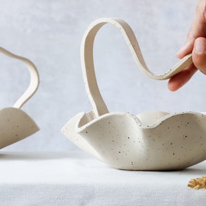 On a table with a white tablecloth is a round basket with a wavy edge. It has a handle of the same material and forms a wave that goes down in the center. One hand tries to lift the basket. Behind it, a part of another basket can be seen.