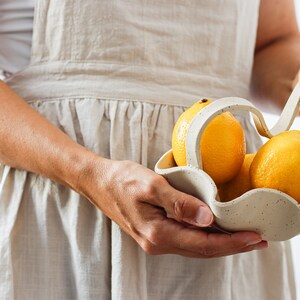 A woman holds almost in profile a beige dotted stoneware wavy basket with a wavy handle. The basket contains three lemons.