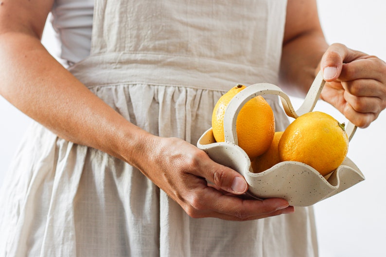 A woman holds almost in profile a beige dotted stoneware wavy basket with a wavy handle. The basket contains three lemons.