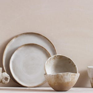 Puede incluir: Una colección de vajilla de cerámica hecha a mano en tonos neutros. El juego incluye dos platos, dos tazas y dos cuencos. Las piezas tienen un acabado rústico y texturizado con una estética natural y orgánica. La paleta de colores es principalmente blanquecina y beige.
