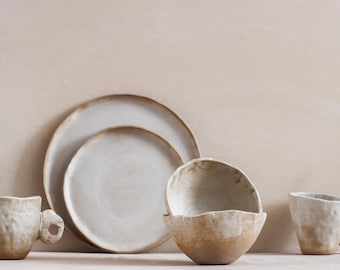 May include: A collection of handmade ceramic tableware in neutral tones. The set includes two plates, two mugs, and two bowls. The pieces have a rustic, textured finish with a natural, organic aesthetic. The color palette is primarily off-white and beige.