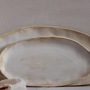 Leaning horizontally against a wall are two oval-shaped, matte white ceramic platters