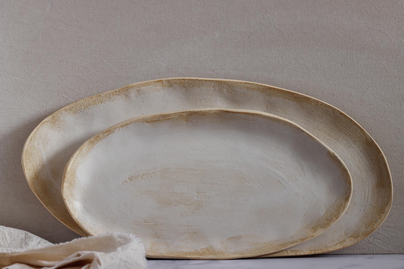 Leaning horizontally against a wall are two oval-shaped, matte white ceramic platters