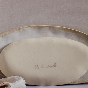 Leaning horizontally against a wall are two oval-shaped, matte white ceramic fountains. The one in front is showing the reverse, with the author's signature.