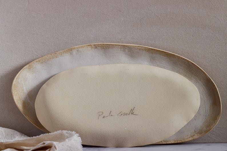 Leaning horizontally against a wall are two oval-shaped, matte white ceramic fountains. The one in front is showing the reverse, with the author's signature.