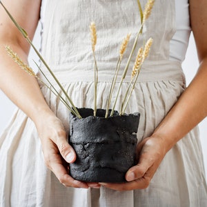 Two hands of a woman in an apron hold a flower pot that is actually a utensil container. It is made from strips of clay cut by hand and randomly joined together. It contains spikes.