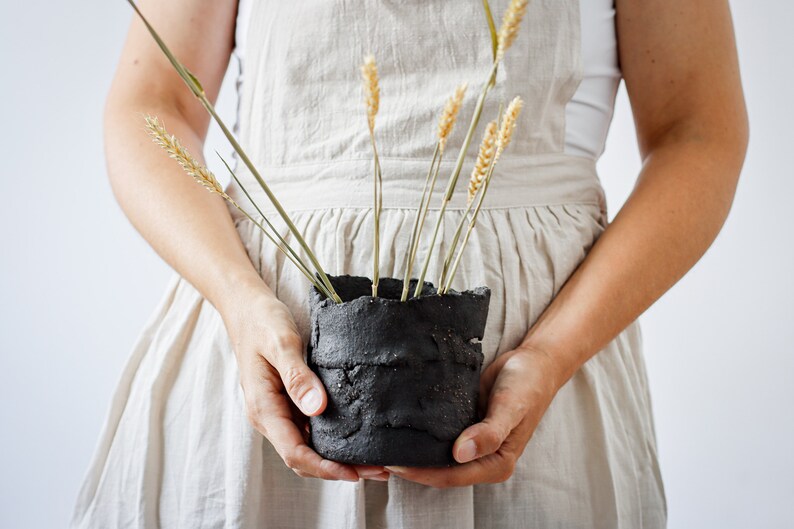 Two hands of a woman in an apron hold a flower pot that is actually a utensil container. It is made from strips of clay cut by hand and randomly joined together. It contains spikes.