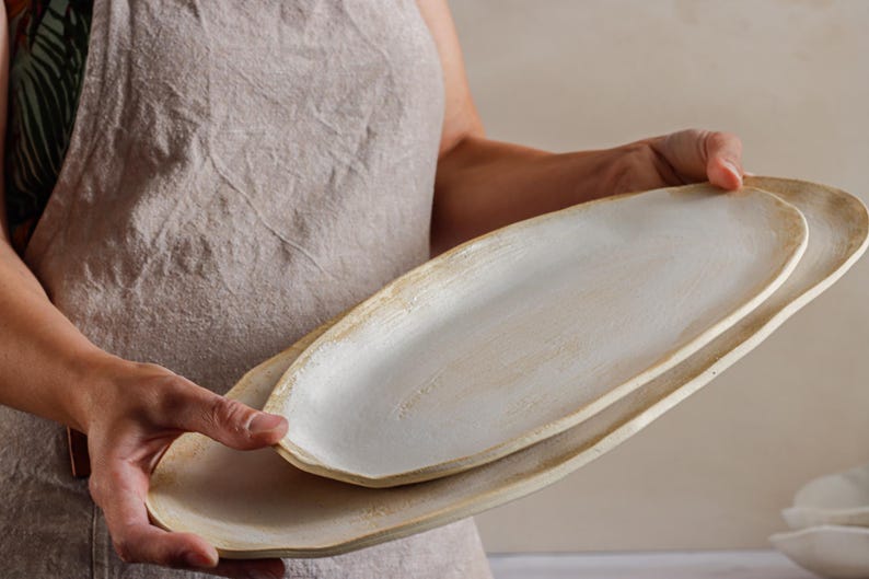 A woman holds in her hands two oval ceramic dishes glazed in matte white. One is smaller than the other