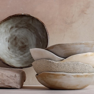 May include: A collection of handmade ceramic bowls in various sizes and textures. The bowls are in shades of beige, cream, and gray, with a rustic, organic aesthetic. One large bowl is placed separately, while others are stacked.