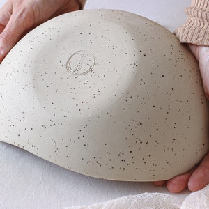 View at 45º. Two hands hold an oval fruit bowl in mottled beige clay. The back of the fruit bowl is visible. It has the artist's seal. The fruit bowl is deep, and a light pink cloth completes the scene.