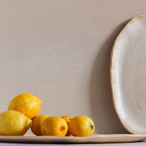 On a table is an oval tray full of lemons. Next to it is another tray with the same shape. It is white matte stoneware