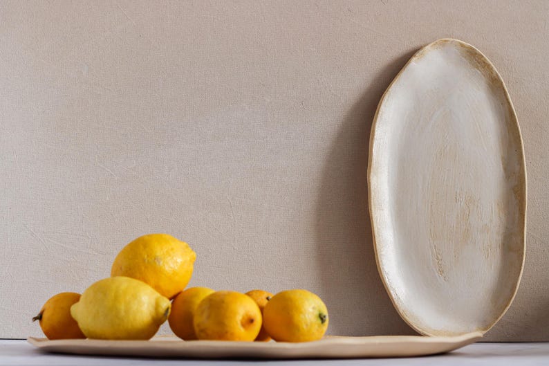 On a table is an oval tray full of lemons. Next to it is another tray with the same shape. It is white matte stoneware