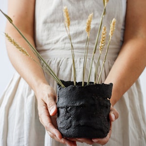 A woman holds a flower pot that is actually a utensil container. It is made from strips of black textured clay cut by hand and randomly joined together. It has some holes. It contains spikes.