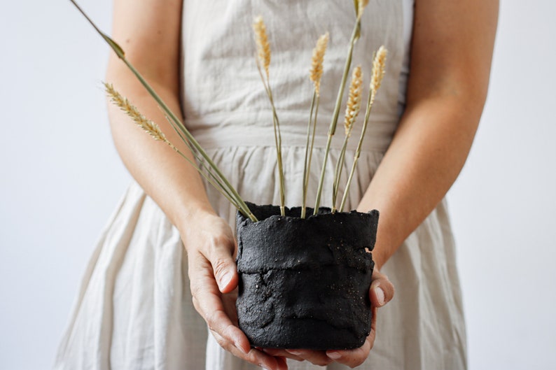 A woman holds a flower pot that is actually a utensil container. It is made from strips of black textured clay cut by hand and randomly joined together. It has some holes. It contains spikes.