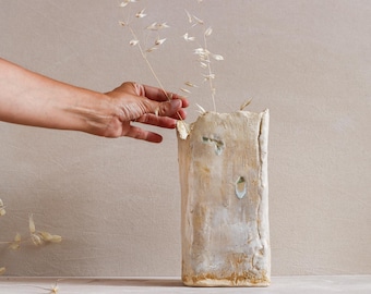 May include: A rectangular, textured, beige ceramic vase with a rough, organic appearance. The vase has several holes and is filled with delicate, dried, light-coloured stems. A hand reaches towards the stems.