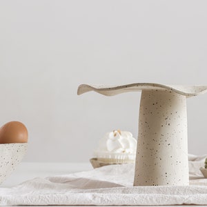 Still life in high-key. On the right is a beige marbled stoneware cake stand. On the left is a part of a bowl of the same material containing eggs. On the table, there is a beige cloth and several sweets.
