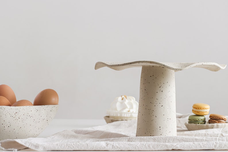 Still life in high-key. On the right is a beige marbled stoneware cake stand. On the left is a part of a bowl of the same material containing eggs. On the table, there is a beige cloth and several sweets.