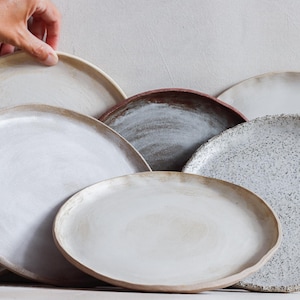 May include: A collection of handmade ceramic plates in various sizes and colors. The plates feature a range of neutral tones, including white, beige, and gray, with some having speckled or textured surfaces. The plates are arranged in a layered composition.