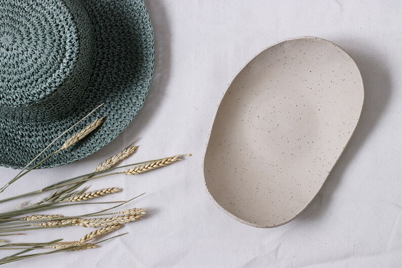 Zenithal view of an oval fruit bowl in mottled beige clay. On the left can be seen a part of a green hat and some ears of corn.