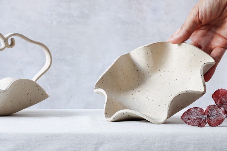 On a table with a white tablecloth is a round bowl with a wavy edge. The inside is visible because a hand is lifting it from the back. To the left is a part of another basket of the same material containing a wave-shaped handle.