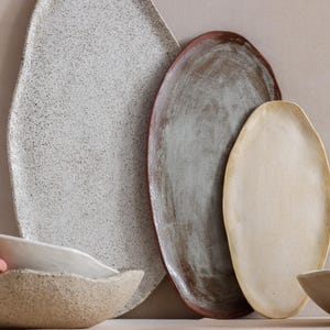 Against a wall, an oval terracotta tray, a larger black and white one to its left, and a smaller beige one to its right. Next to it, on a table, a hand lifts a stoneware ceramic bowl resting on top of another of the same style.