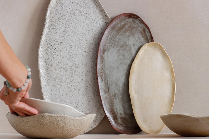 Against a wall, an oval terracotta tray, a larger black and white one to its left, and a smaller beige one to its right. Next to it, on a table, a hand lifts a stoneware ceramic bowl resting on top of another of the same style.