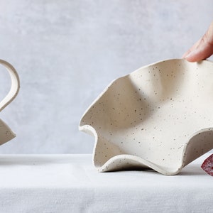 On a table is a round dotted ceramic bowl with a wavy edge. The inside is visible because a hand is lifting it from the back. To the left is a part of another basket of the same material containing a wave-shaped handle.