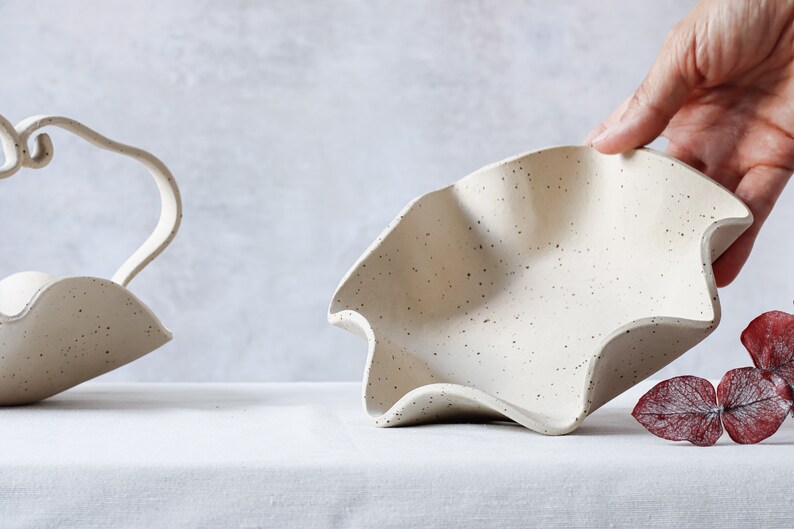 On a table is a round dotted ceramic bowl with a wavy edge. The inside is visible because a hand is lifting it from the back. To the left is a part of another basket of the same material containing a wave-shaped handle.