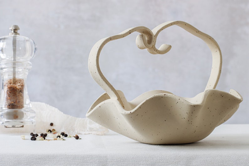 Front view of a mottled beige ceramic basket on a table with a white tablecloth. It has a spiral handle in the center. A glass salt grinder can be seen behind it.