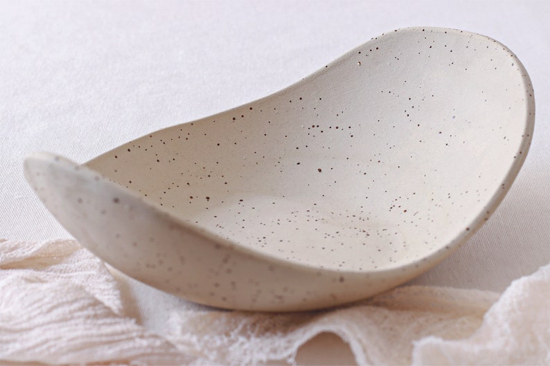 Front view of a fruit bowl seen diagonally. It is made of mottled beige clay and is deep and oval in shape.