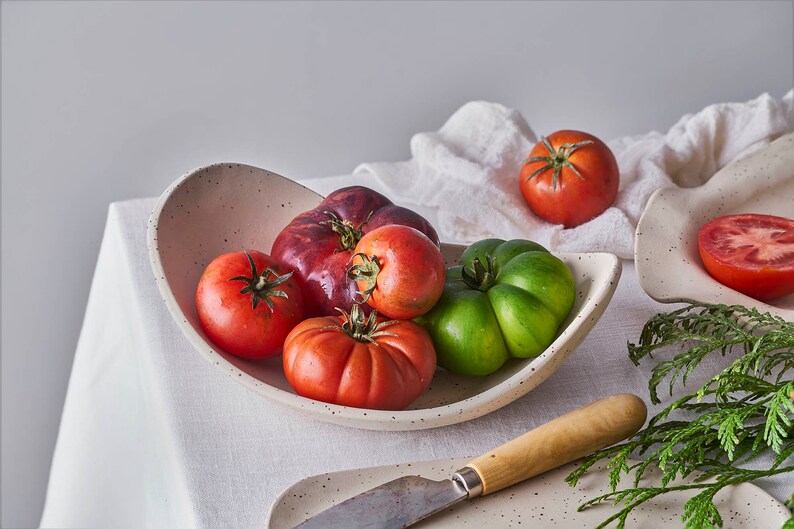 A beige-mottled oval fruit bowl holds five tomatoes. Still life photo. On the white tablecloth table is also a knife, more tomatoes, herbs, and a cloth.