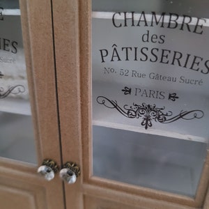 May include: A wooden cabinet with glass doors. The doors have ornate black lettering that reads "Chambre des P&acirc;tisseries No. 52 Rue G&acirc;teau Sucr&eacute; PARIS".