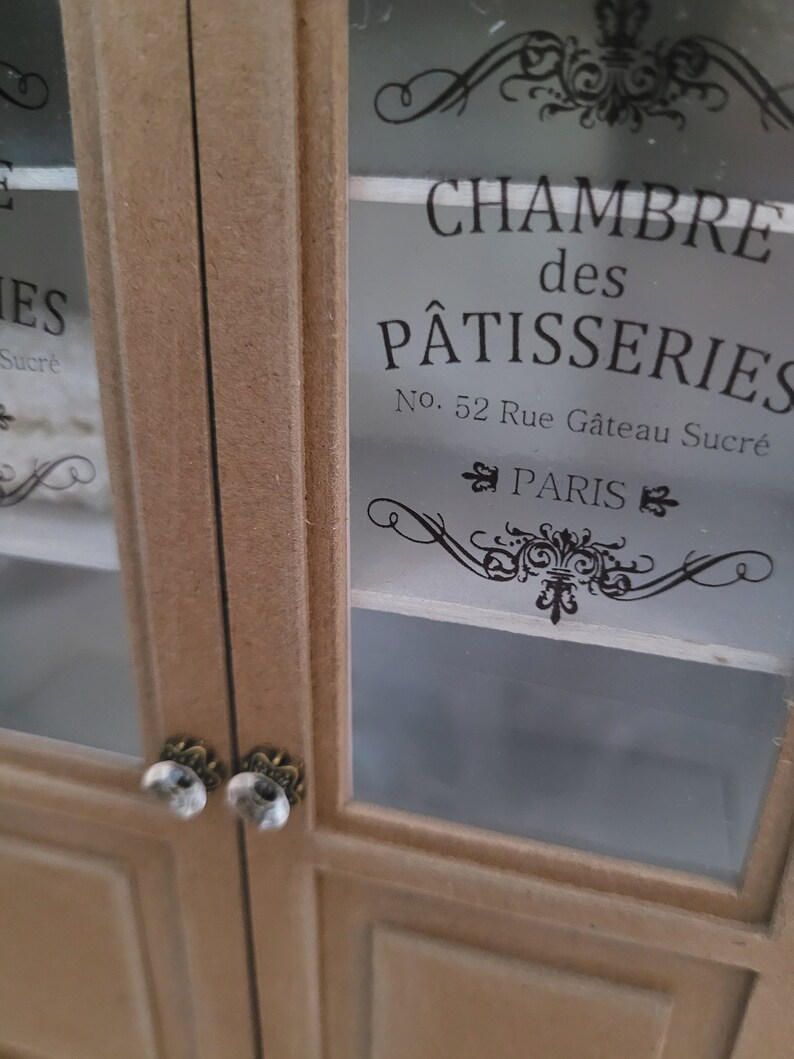 May include: A wooden cabinet with glass doors. The doors have ornate black lettering that reads "Chambre des P&acirc;tisseries No. 52 Rue G&acirc;teau Sucr&eacute; PARIS".