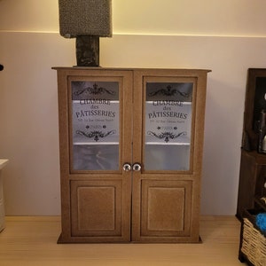 May include: A brown wooden cabinet with two glass doors. The doors have a decorative design and the text "CHAMBRE des P&Acirc;TISSERIES No. 52 Rue G&acirc;teau Sucr&eacute; PARIS" on each door.