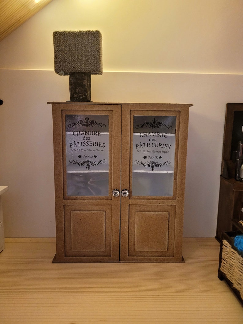 May include: A brown wooden cabinet with two glass doors. The doors have a decorative design and the text "CHAMBRE des P&Acirc;TISSERIES No. 52 Rue G&acirc;teau Sucr&eacute; PARIS" on each door.