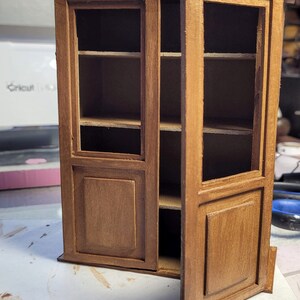 May include: A wooden cabinet with glass doors and shelves. The cabinet is painted a light brown color and has a distressed finish. The doors are open and the shelves are empty.