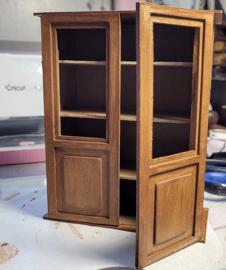 May include: A wooden cabinet with glass doors and shelves. The cabinet is painted a light brown color and has a distressed finish. The doors are open and the shelves are empty.
