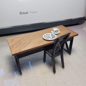 May include: A miniature wooden table with a black base and a wood top. A black chair sits next to the table. A white teacup and saucer with a black floral design sits on a white plate with a black floral design on the table.