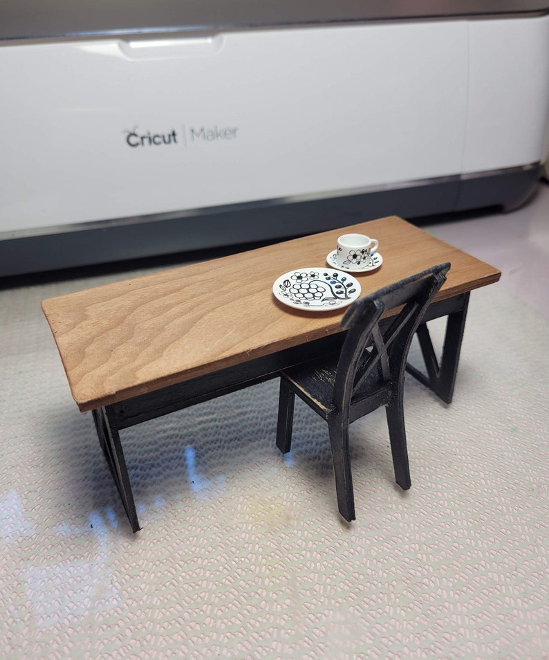 May include: A miniature wooden table with a black base and a wood top. A black chair sits next to the table. A white teacup and saucer with a black floral design sits on a white plate with a black floral design on the table.