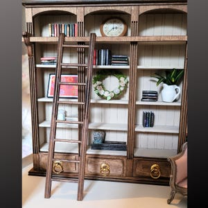 May include: A miniature wooden bookcase with three vertical sections, arched tops, and three bottom drawers with gold ring pulls. A tall wooden ladder leans against the shelves, which contain miniature books, a round clock, a floral wreath, and a white pitcher.