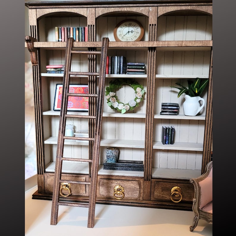 May include: A miniature wooden bookcase with three vertical sections, arched tops, and three bottom drawers with gold ring pulls. A tall wooden ladder leans against the shelves, which contain miniature books, a round clock, a floral wreath, and a white pitcher.