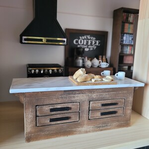 May include: Miniature kitchen scene with a wooden island featuring a marble countertop and two drawers. A black range hood and stove are visible. A chalkboard sign reads "Brewed Coffee served daily". Bread and coffee cups are on the counter.