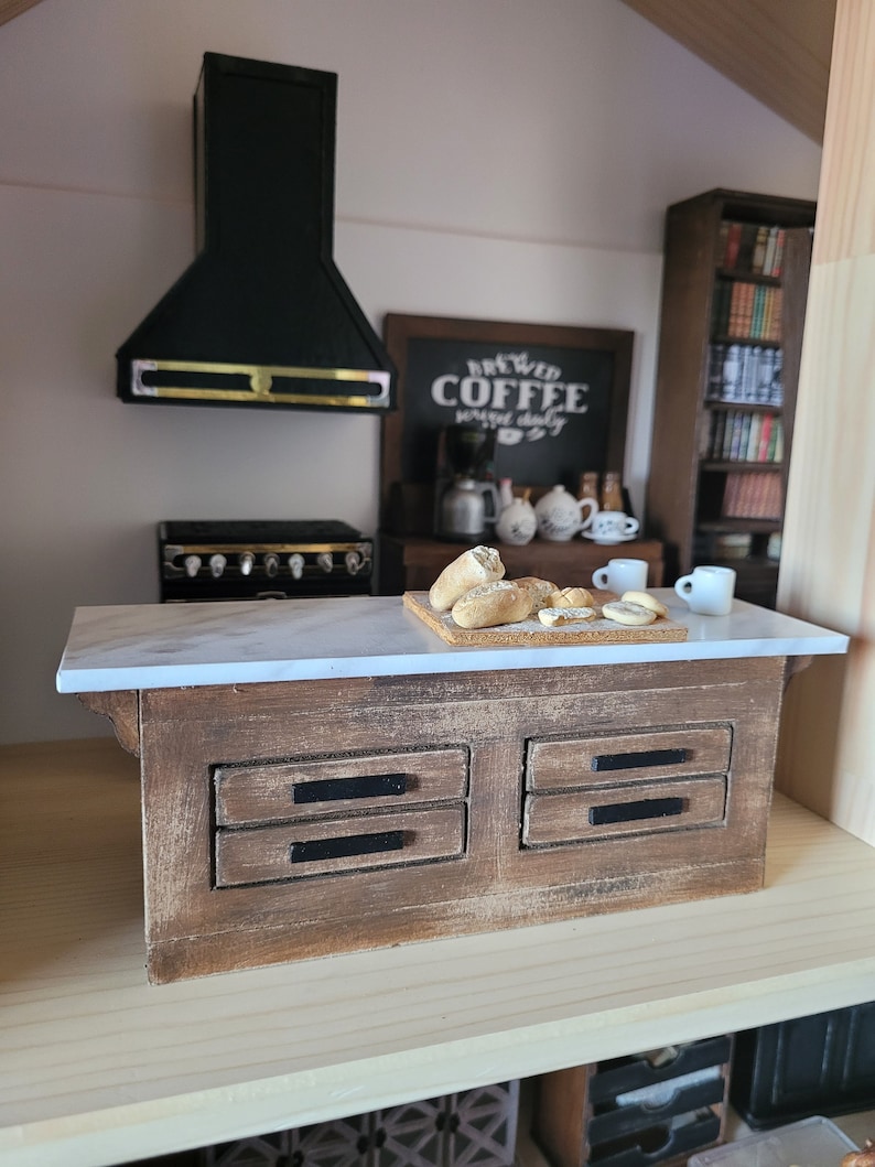 May include: Miniature kitchen scene with a wooden island featuring a marble countertop and two drawers. A black range hood and stove are visible. A chalkboard sign reads "Brewed Coffee served daily". Bread and coffee cups are on the counter.