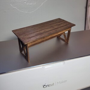 May include: A miniature wooden table with a brown stain finish. The table has a rectangular top with a simple design and a base with an X-shaped support.